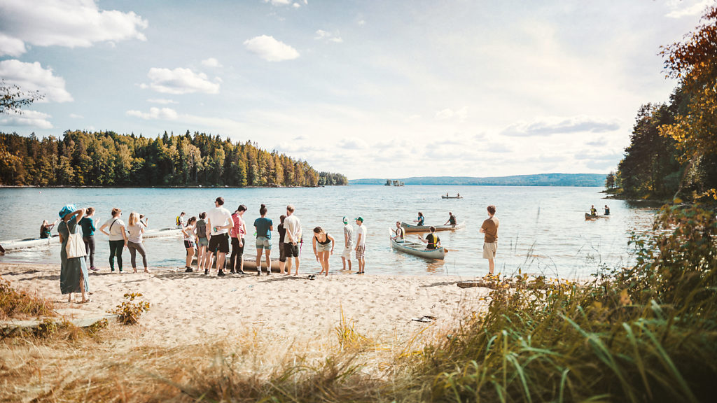 A group of people standing next to a beach preparing for a canoe trip.