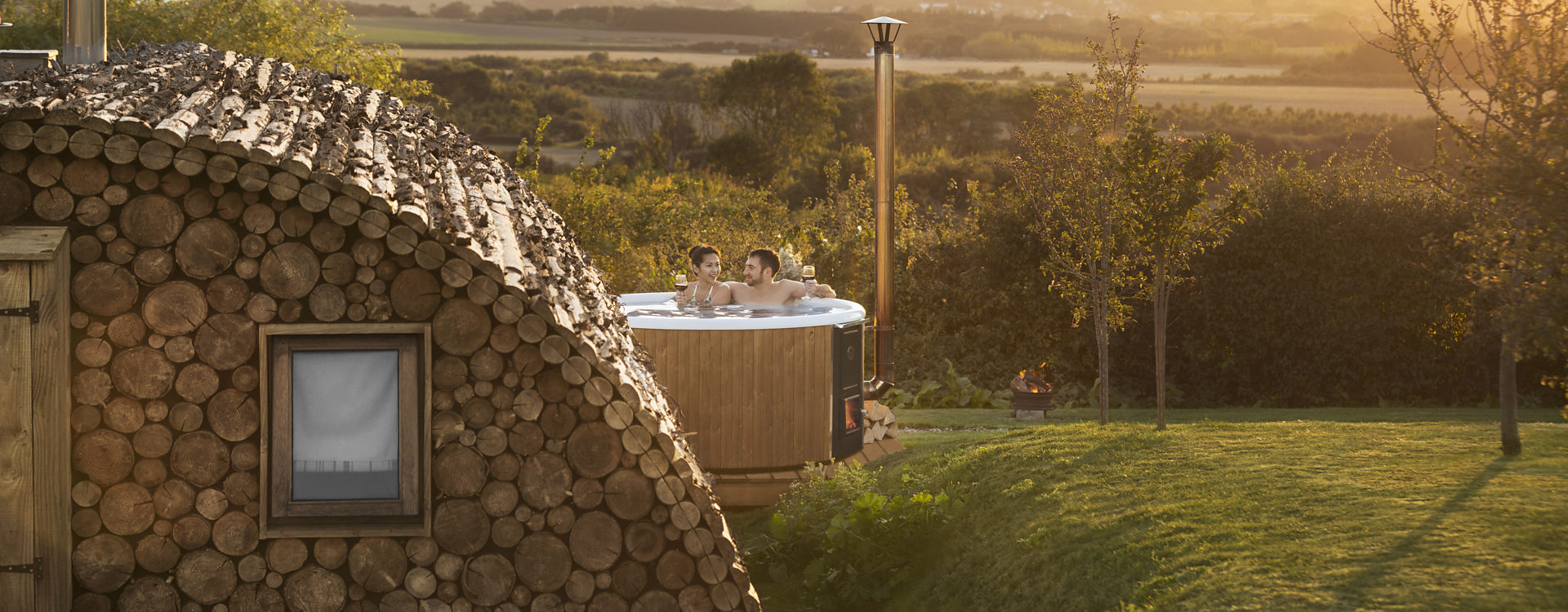 A sunset picture of a couple sitting in a Skargards Regal that is placed next to a wooden house and surrounded by gras.